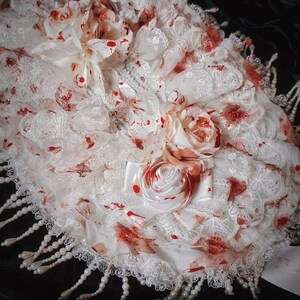 May include: A white lace decorative item with red splatters and floral accents. The item features a layered design with a scalloped edge and is adorned with faux pearls. The overall aesthetic is gothic and macabre.