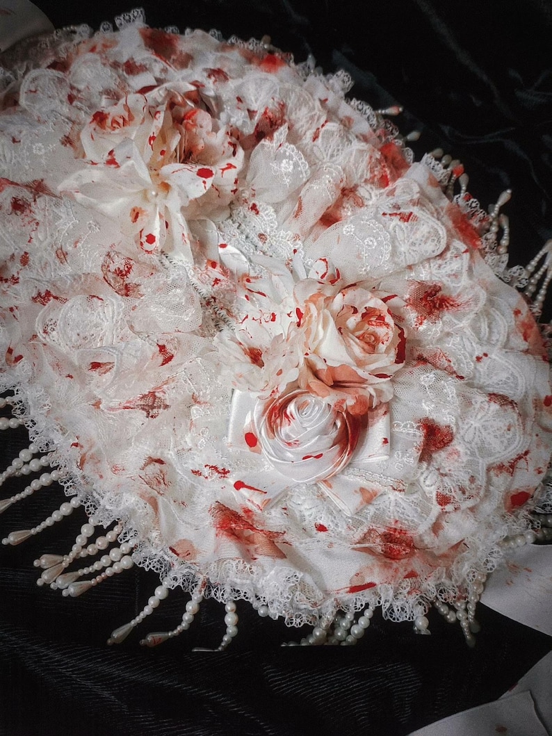 May include: A white lace decorative item with red splatters and floral accents. The item features a layered design with a scalloped edge and is adorned with faux pearls. The overall aesthetic is gothic and macabre.