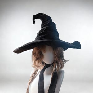 以下が含まれることがあります： 幅広のつばと尖ったクラウンが付いた黒い魔女の帽子。帽子は柔らかく暗い生地で作られています。帽子は、明るい茶色の髪と黒いリボンが付いたマネキンの頭に飾られています。