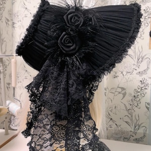 Op de afbeelding: Zwarte bonnet met geplooid stof, versierd met twee stoffen rozen en veren. De bonnet heeft een kanten sluier. De hoed is in gotische of Victoriaanse stijl.