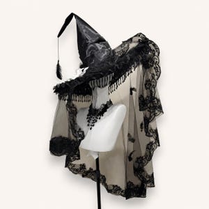 May include: A black pointed hat with a wide brim, featuring intricate lace trim, hanging fringe, and a long sheer veil with floral lace edges. A matching black lace choker with dangling teardrop accents is displayed on a white mannequin neck.