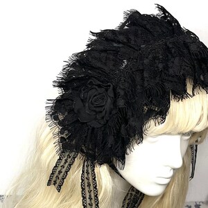 May include: A black lace headpiece featuring layered, ruffled fabric with frayed edges and a central fabric rose. The accessory includes two dangling lace ribbons with a delicate floral pattern, designed to be worn over long blonde hair.