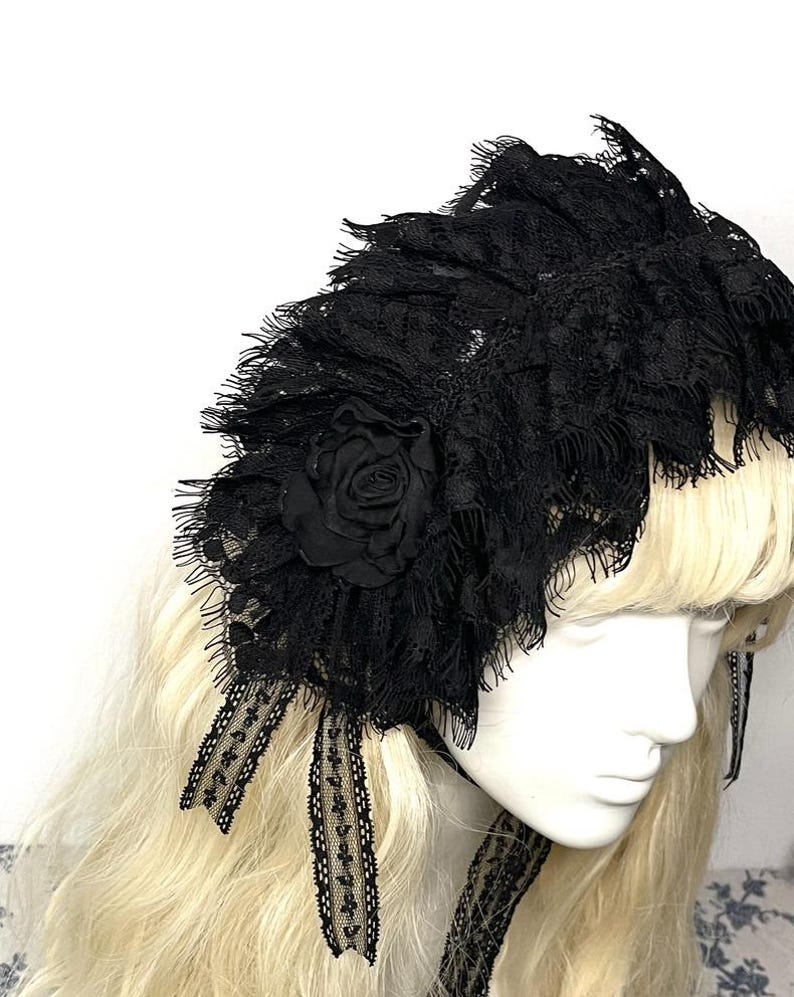 May include: A black lace headpiece featuring layered, ruffled fabric with frayed edges and a central fabric rose. The accessory includes two dangling lace ribbons with a delicate floral pattern, designed to be worn over long blonde hair.