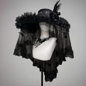 May include: A black, wide-brimmed hat with floral and feather detailing, displayed on a mannequin. The hat has a lace veil and beaded fringe. A black lace necklace is also present.