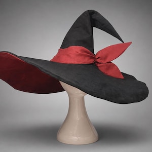 Peut inclure: Un chapeau pointu noir doté d'un large bord et d'une bande en tissu rouge foncé nouée en nœud à la base de la calotte. Le dessous du bord est également doublé de tissu rouge foncé, contrastant avec l'extérieur noir du cône haut et incurvé.