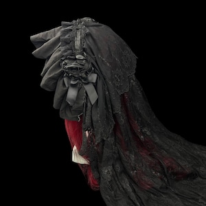 May include: A black bonnet featuring layered fabric ruffles, a decorative fabric rose with a small bow, and a long, intricate lace veil that drapes downward. The headpiece is shown against a black background, highlighting the dark textile details.