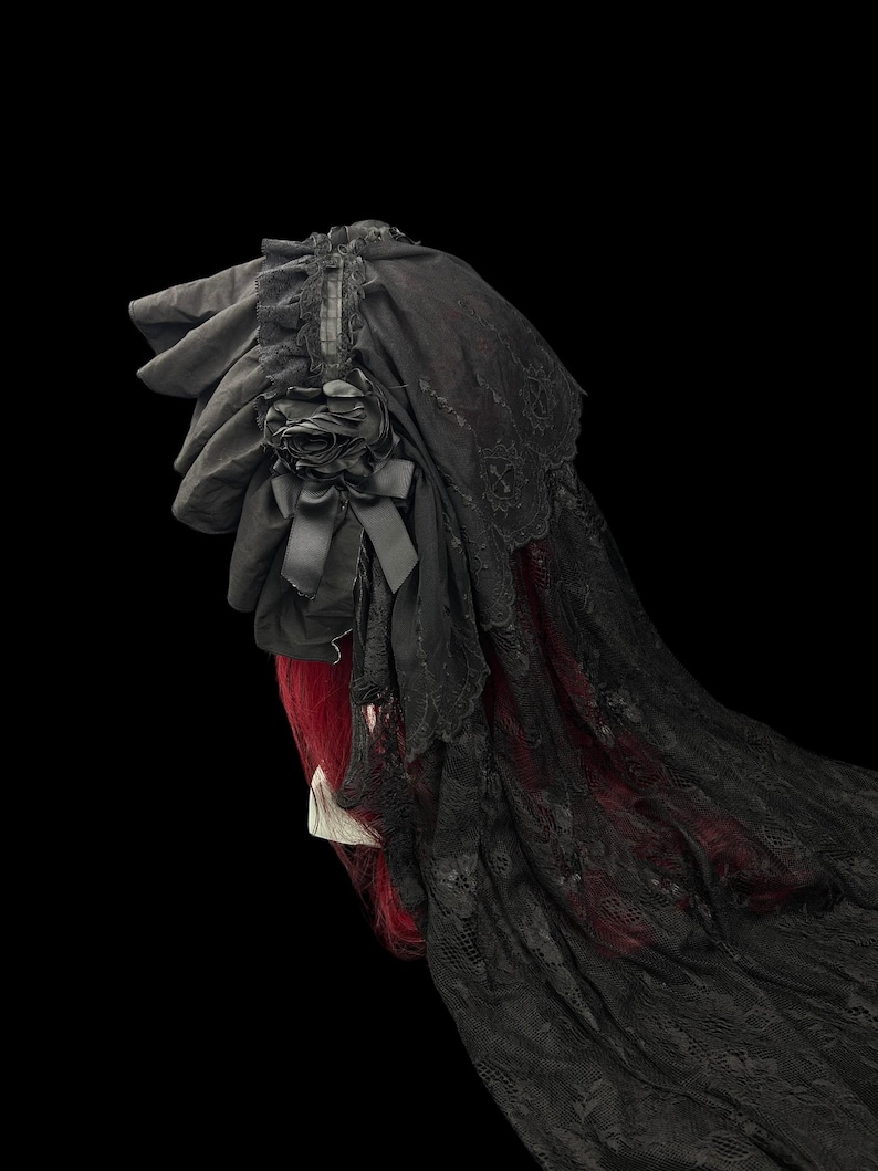 May include: A black bonnet featuring layered fabric ruffles, a decorative fabric rose with a small bow, and a long, intricate lace veil that drapes downward. The headpiece is shown against a black background, highlighting the dark textile details.