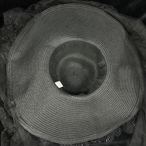 Puede incluir: Un sombrero negro de ala ancha con textura tejida circular y copa profunda. El sombrero est&aacute; adornado con una capa de tela de tul negro transparente que se extiende m&aacute;s all&aacute; del ala, acentuada con peque&ntilde;as cuentas negras colgantes en el borde.