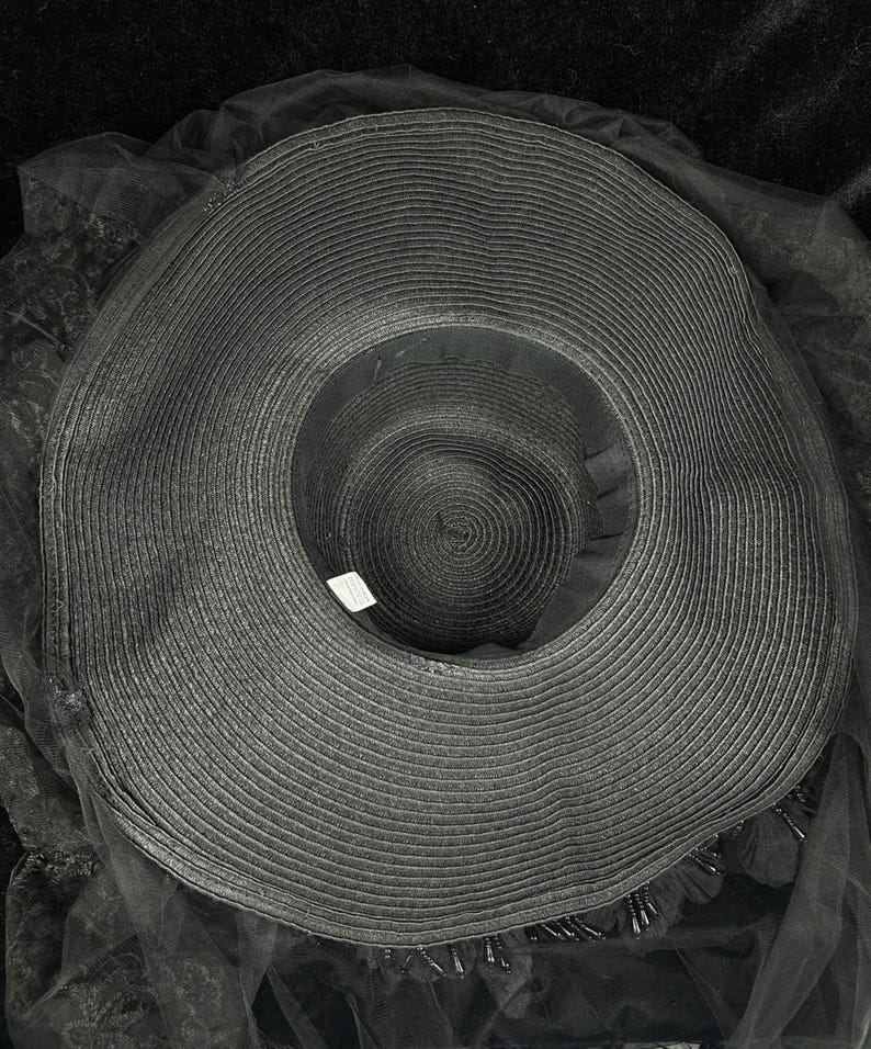 Puede incluir: Un sombrero negro de ala ancha con textura tejida circular y copa profunda. El sombrero est&aacute; adornado con una capa de tela de tul negro transparente que se extiende m&aacute;s all&aacute; del ala, acentuada con peque&ntilde;as cuentas negras colgantes en el borde.