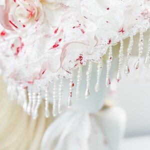 May include: A white bonnet with red accents and a floral design. The bonnet features layers of lace and fabric, adorned with dangling pearl beads. The design includes red splatters and a large flower, creating a unique and decorative headpiece.