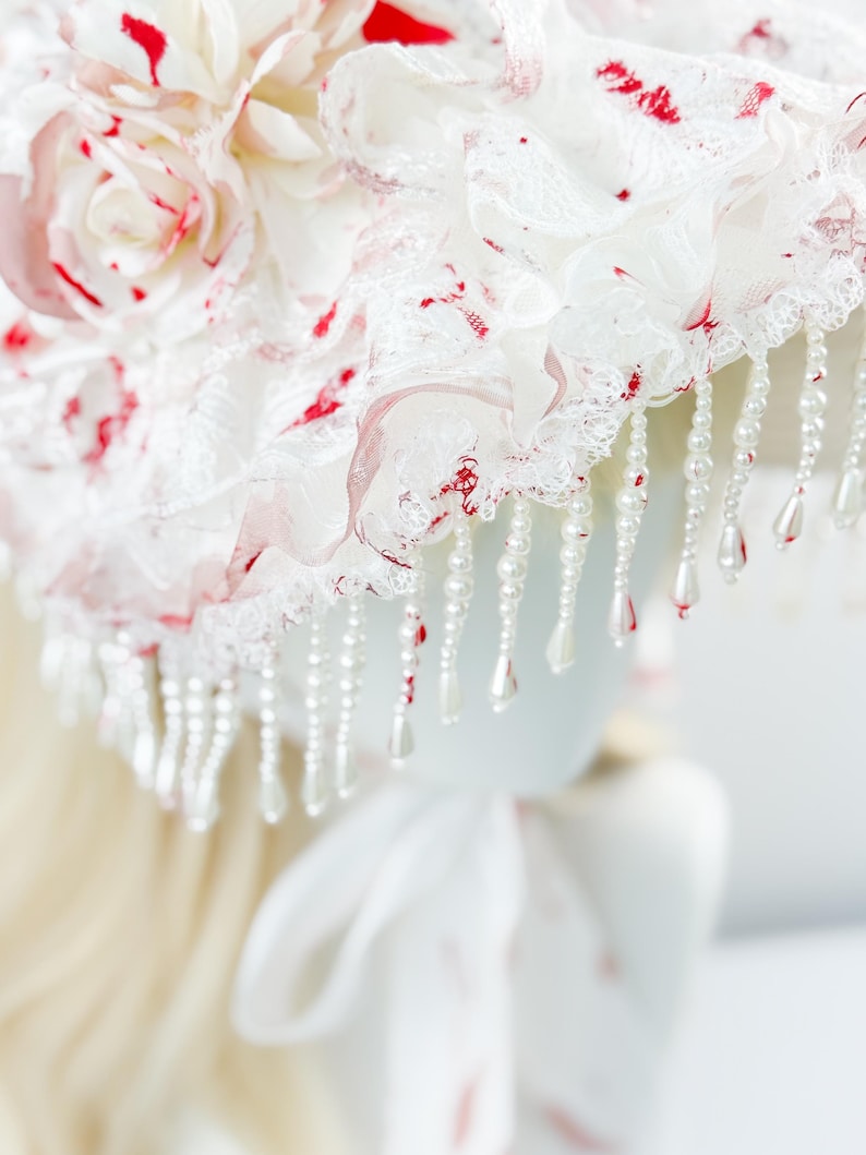 May include: A white bonnet with red accents and a floral design. The bonnet features layers of lace and fabric, adorned with dangling pearl beads. The design includes red splatters and a large flower, creating a unique and decorative headpiece.