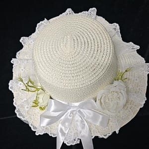 May include: A cream-colored straw hat featuring a wide brim adorned with white lace trim. The hat is decorated with two white fabric roses, green sprigs, and a white satin ribbon tied into a bow at the front, with two long ribbon tails hanging down.