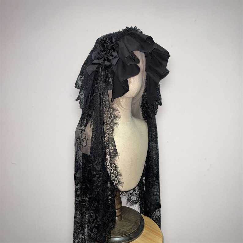 May include: A black lace head covering featuring a ruffled fabric hood and a decorative fabric flower accent. The intricate lace pattern extends down the sides, creating a draped effect over the shoulders. The headpiece is displayed on a mannequin.