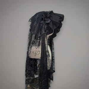 May include: A black headpiece featuring a ruffled fabric brim and a decorative fabric rose. The accessory includes a long, intricate lace veil with a floral pattern that drapes downward, covering the sides and back of the headpiece structure.