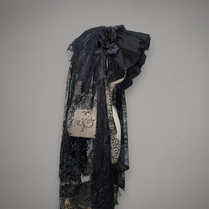 May include: A black headpiece featuring a ruffled fabric brim and a decorative fabric rose. The accessory includes a long, intricate lace veil with a floral pattern that drapes downward, covering the sides and back of the headpiece structure.