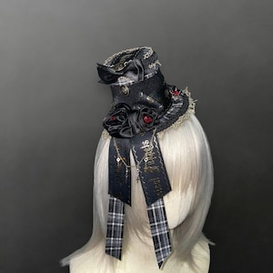 May include: A black top hat with decorative elements. The hat features black satin roses with red accents, a black ribbon with gold text, and a plaid ribbon. The hat is trimmed with lace and has a small bow.