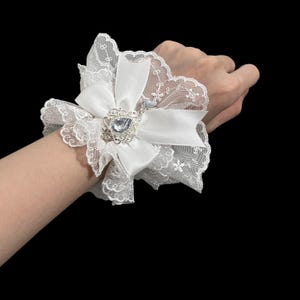 May include: White lace wrist cuff with a satin bow and a silver-toned embellishment featuring a large, clear gemstone. The cuff has a delicate, scalloped edge and is designed to be worn as a fashion accessory.