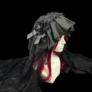 May include: A black bonnet featuring ruffled fabric, a decorative fabric rose, and long lace panels that drape down the sides. The headpiece is displayed on a mannequin head with long red hair, showcasing the intricate lace texture and layered design.