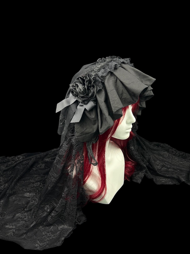 May include: A black bonnet featuring ruffled fabric, a decorative fabric rose, and long lace panels that drape down the sides. The headpiece is displayed on a mannequin head with long red hair, showcasing the intricate lace texture and layered design.