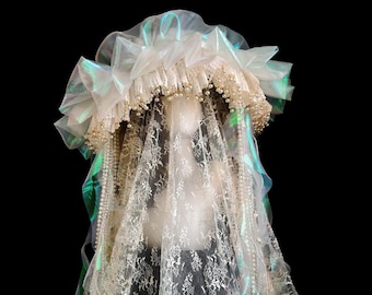 Può includere: Un copricapo con una corona di volant in organza plissettata iridescente e un lungo velo in pizzo floreale bianco. La base è decorata con numerose piccole perle artificiali color crema che scendono lungo i lati, creando un aspetto stratificato.