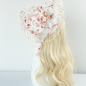 May include: A white headpiece with a floral design and red accents. The headpiece features lace, faux pearls, and a cascade of beaded fringe. The design includes fabric roses and red splatters, creating a unique aesthetic.