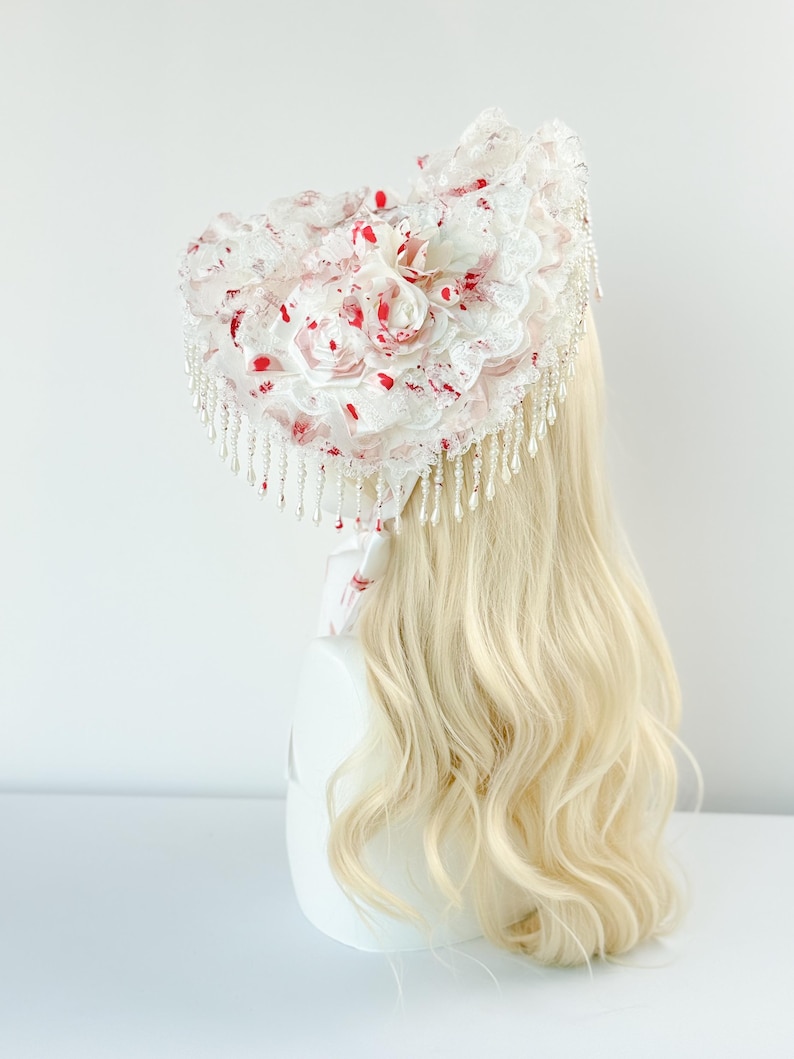 May include: A white headpiece with a floral design and red accents. The headpiece features lace, faux pearls, and a cascade of beaded fringe. The design includes fabric roses and red splatters, creating a unique aesthetic.
