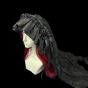 May include: A black headpiece featuring a ruffled fabric brim and a long, sheer lace veil. The headpiece is adorned with a fabric rose and ribbon detail on the side, displayed on a mannequin head with long red hair against a black background.