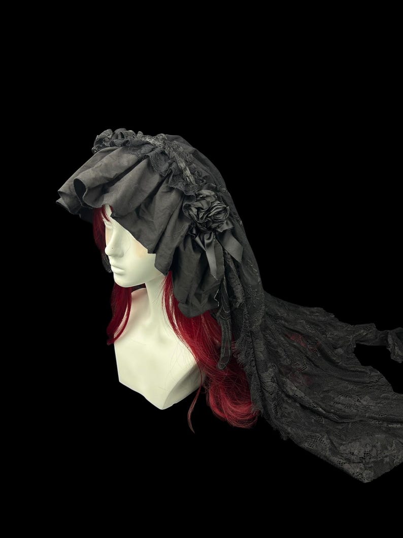 May include: A black headpiece featuring a ruffled fabric brim and a long, sheer lace veil. The headpiece is adorned with a fabric rose and ribbon detail on the side, displayed on a mannequin head with long red hair against a black background.