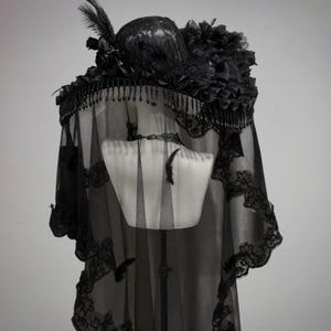 Puede incluir: Un sombrero negro con ala ancha adornado con cuentas colgantes, encaje y plumas. Un velo negro largo y transparente con un intrincado borde de encaje cuelga del ala, cubriendo los hombros. Se lleva una gargantilla de encaje negro a juego.