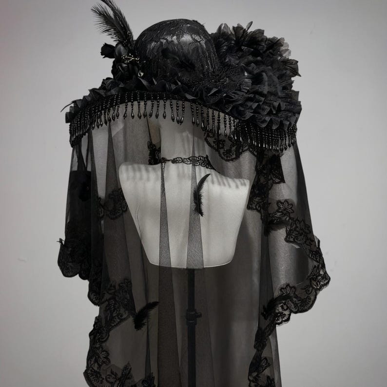 Puede incluir: Un sombrero negro con ala ancha adornado con cuentas colgantes, encaje y plumas. Un velo negro largo y transparente con un intrincado borde de encaje cuelga del ala, cubriendo los hombros. Se lleva una gargantilla de encaje negro a juego.