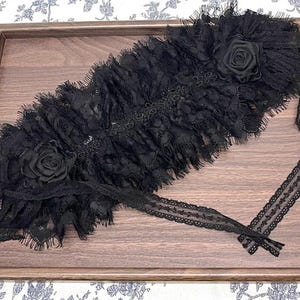 May include: A black lace garter featuring a layered, ruffled design with two fabric roses attached at opposite ends. The accessory is constructed from intricate, sheer black lace with frayed edges and includes long, thin lace ties for fastening.