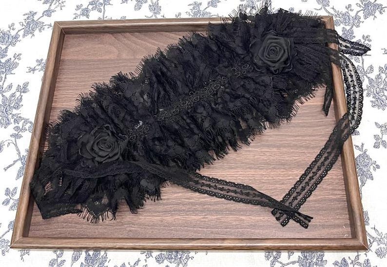 May include: A black lace garter featuring a layered, ruffled design with two fabric roses attached at opposite ends. The accessory is constructed from intricate, sheer black lace with frayed edges and includes long, thin lace ties for fastening.