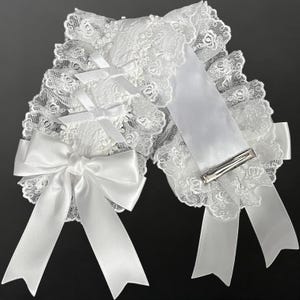 May include: A pair of white hair accessories featuring intricate floral lace trim, layered satin ribbons, and small decorative bows. Each piece is finished with a silver-toned metal alligator clip attached to the back for secure placement in hair.