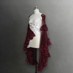 Op de afbeelding: Een witte strapless korsettop met een delicaat bloemenpatroon, gecombineerd met een transparant, diepbordeauxrood mesh bolero. Het bolero heeft lange, zwierige mouwen versierd met meerdere lagen ruches en een afwerking van kanten randen.