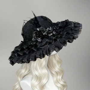 May include: A black, wide-brimmed hat with a ruffled lace brim and decorative black roses. The hat features a high crown and is adorned with feathers. The hat is displayed on a mannequin head with long, wavy, blonde hair.