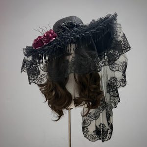 Puede incluir: Un sombrero negro de ala ancha, adornado con encaje negro, flecos y un velo. El sombrero presenta un racimo de flores rojo oscuro y se exhibe en una cabeza de maniquí. El diseño es gótico y elegante.