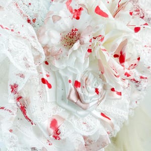 May include: Close-up of a white fabric headpiece with red splatter details. The headpiece features layers of lace, fabric flowers, and a small satin rose. Pearl beads hang from the bottom edge, adding a delicate touch to the design.