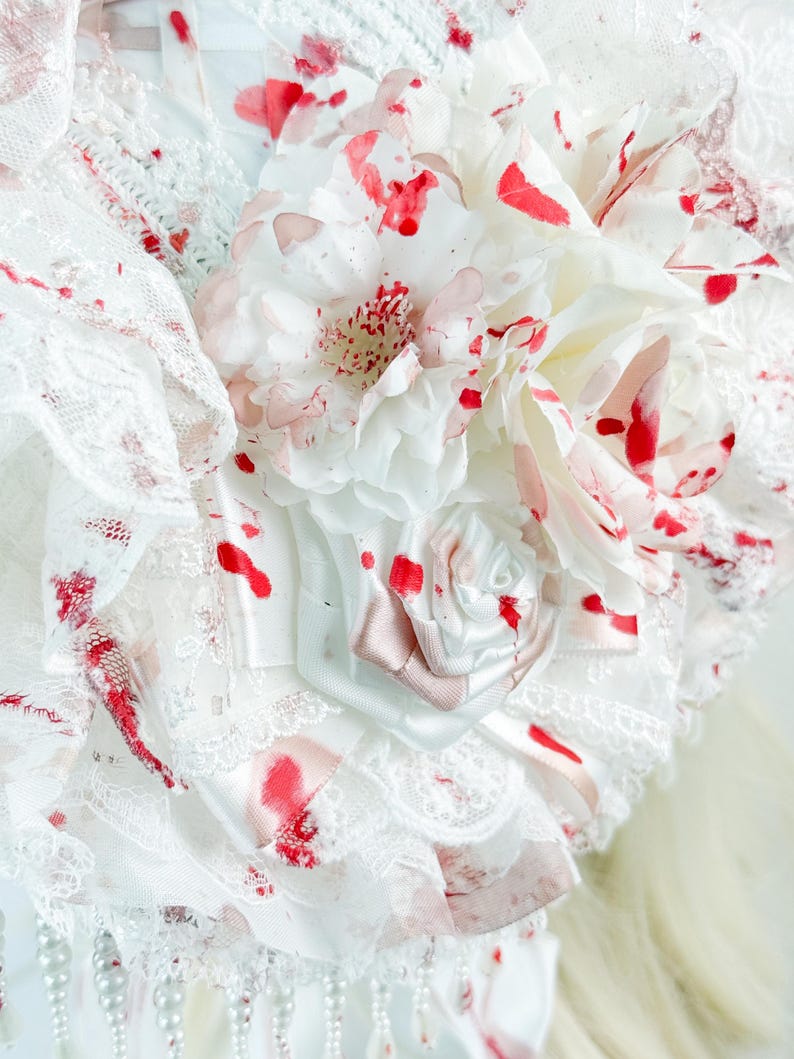 May include: Close-up of a white fabric headpiece with red splatter details. The headpiece features layers of lace, fabric flowers, and a small satin rose. Pearl beads hang from the bottom edge, adding a delicate touch to the design.