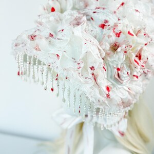 May include: A white headpiece with red accents, featuring layers of lace, fabric flowers, and dangling pearl beads. The headpiece has a bow and ribbons at the back. The design is ornate and detailed, suitable for special occasions.