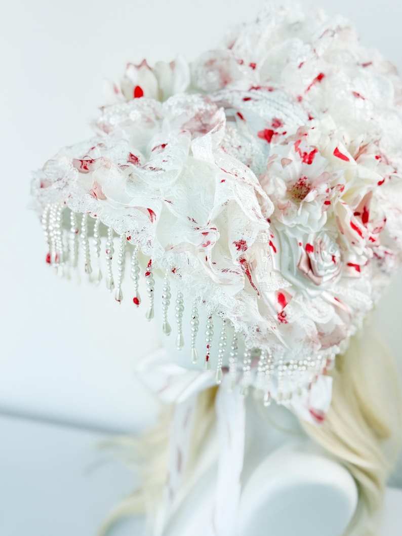 May include: A white headpiece with red accents, featuring layers of lace, fabric flowers, and dangling pearl beads. The headpiece has a bow and ribbons at the back. The design is ornate and detailed, suitable for special occasions.