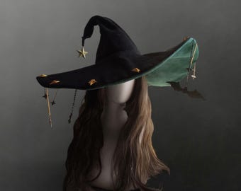 May include: A black witch hat with a green underside, featuring gold-toned star and pyramid-shaped studs along the brim. The hat is adorned with dangling gold chains, small star charms, and a black bat silhouette charm hanging from the brim.