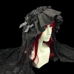 May include: A black bonnet featuring a ruffled brim, a fabric rose accent, and long lace veiling that drapes downwards. The headpiece is displayed on a mannequin head with red hair, showcasing the intricate lace pattern and layered fabric construction.