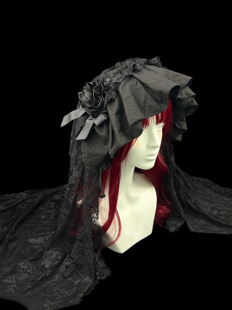 May include: A black bonnet featuring a ruffled brim, a fabric rose accent, and long lace veiling that drapes downwards. The headpiece is displayed on a mannequin head with red hair, showcasing the intricate lace pattern and layered fabric construction.