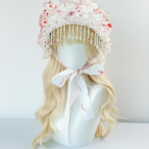 May include: A white bonnet with a floral design and beaded fringe. The bonnet features a layered construction with pink and red accents. A white ribbon with pink markings is tied around the neck of the mannequin head.