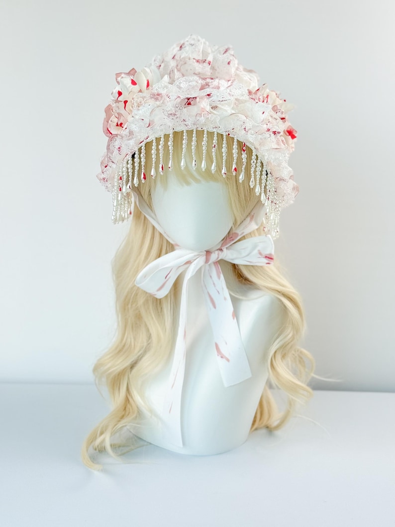 May include: A white bonnet with a floral design and beaded fringe. The bonnet features a layered construction with pink and red accents. A white ribbon with pink markings is tied around the neck of the mannequin head.