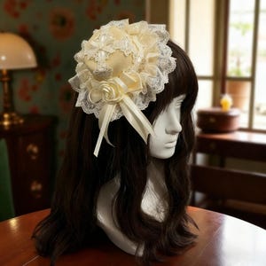 May include: A cream-coloured headpiece with a floral design, lace trim, and a satin bow. The headpiece is adorned with pearls and a fabric rose. The item is displayed on a mannequin head with long, dark brown hair.