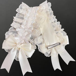 May include: Cream-coloured lace collar with a large satin bow and ribbon details. The collar has delicate lace trim and small bows, with a silver barrette clip for attachment. The design is a mix of textures and patterns.