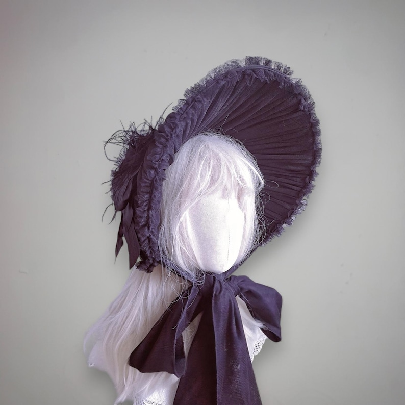 Op de afbeelding: Een donkerpaarse bonnet met een brede rand, versierd met veren en een gerimpelde rand. De hoed heeft een grote strik en wordt getoond op een mannequinhoofd met wit haar. De bonnet is een vintage stijl.