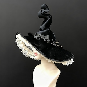 May include: A black velvet witch hat with a wide brim, adorned with a white lace trim and black satin bows. The hat features a tall, spiraling point. The hat is displayed on a mannequin head.