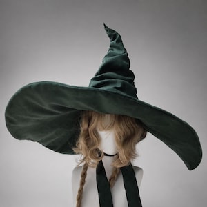May include: A dark green velvet witch hat with a wide, floppy brim and a tall, pointed, slouchy crown. The hat features a soft, textured fabric finish and two long, matching green velvet ribbons hanging down from the base of the crown.